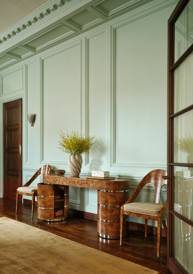 Elegant hallway with mint green paneled walls, a wooden console table, two curved wooden chairs with upholstery, and a vase of yellow flowers on the table.
