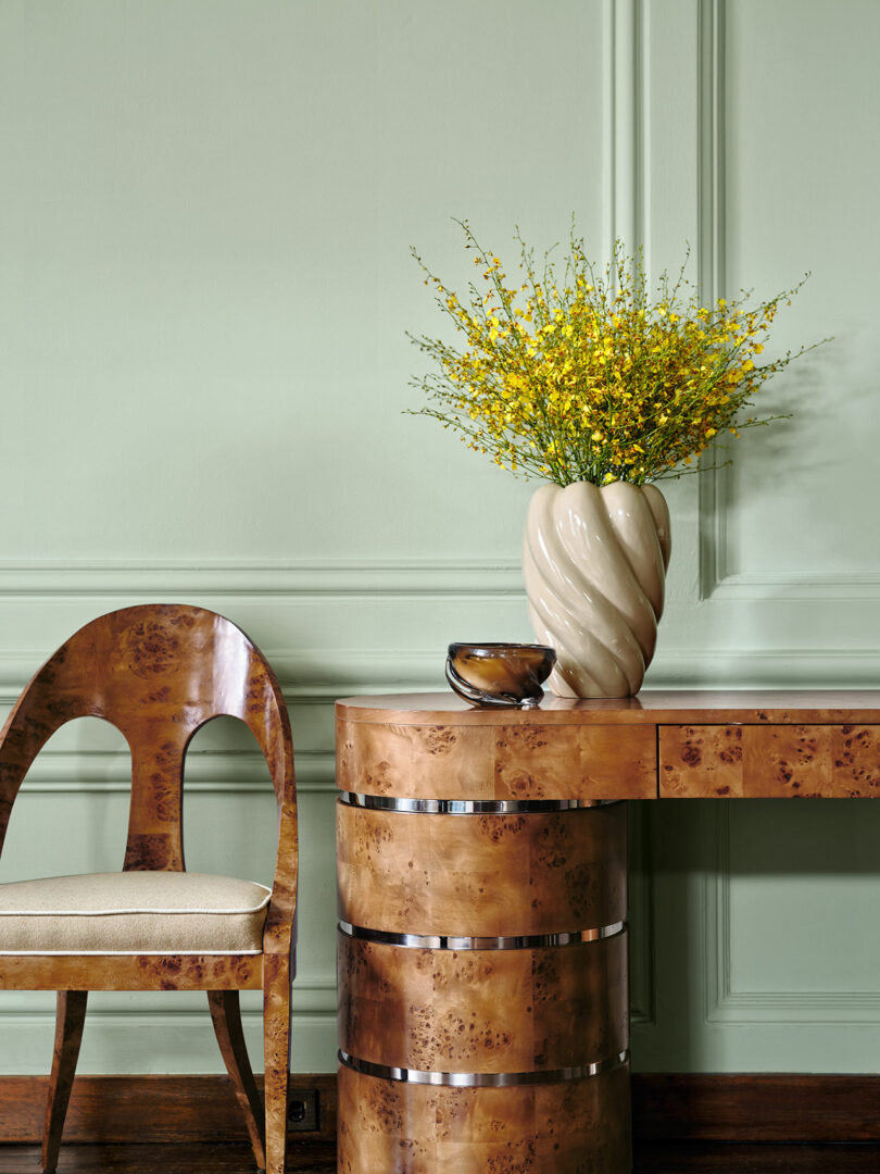 A wooden chair and curved desk sit against a green wall; a cream vase with yellow flowers and a small bowl rest on the desk.