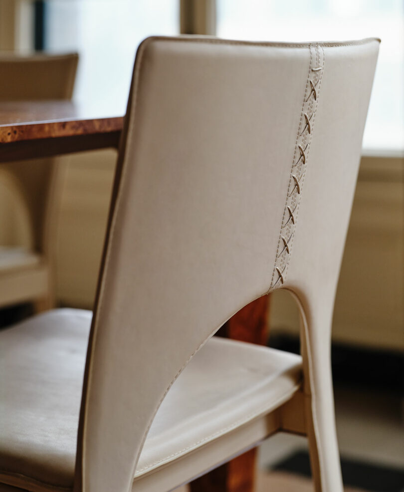 Close-up of a beige upholstered chair with decorative stitching on the backrest, positioned next to a wooden table in a well-lit room.