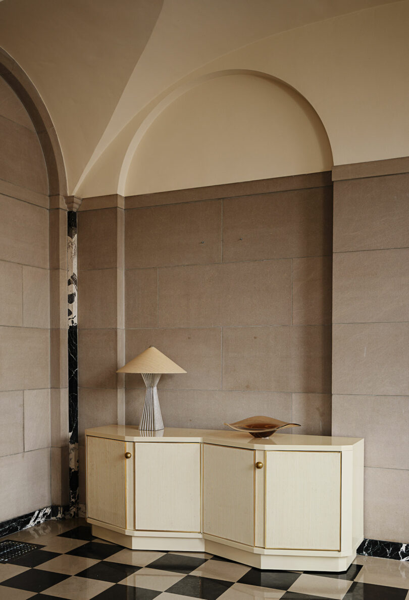 A cream-colored sideboard with gold handles stands against a stone wall, topped with a sculptural lamp and a decorative bowl on a black and white checkered floor.