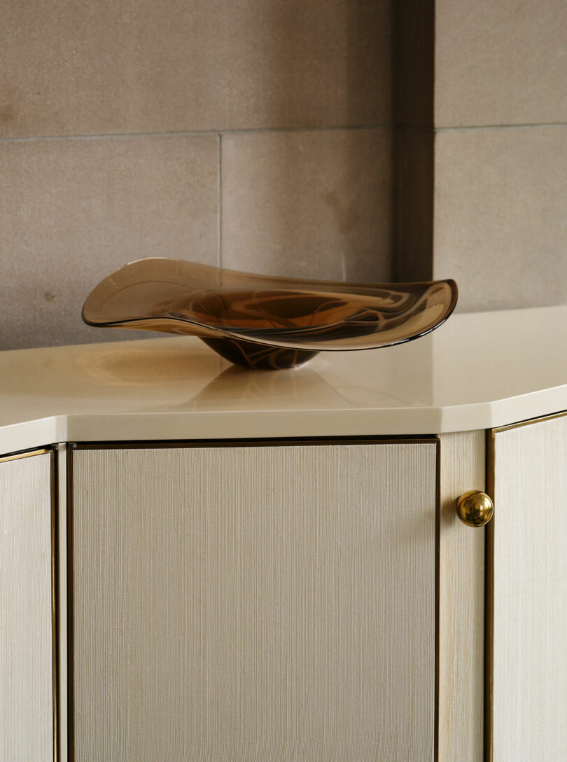 A wavy, translucent brown glass bowl sits on a cream-colored cabinet with gold handles against a beige tiled wall.