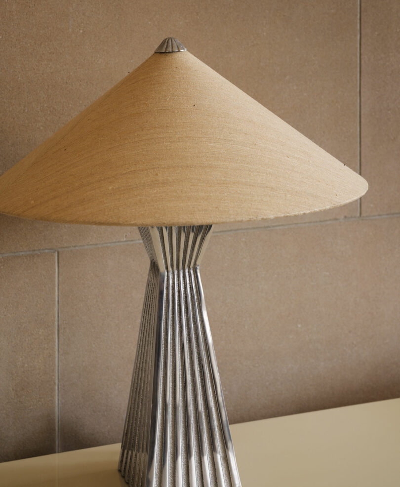 A table lamp with a pleated metallic base and a wide, cone-shaped beige lampshade sits on a cream-colored surface against a stone tile wall.