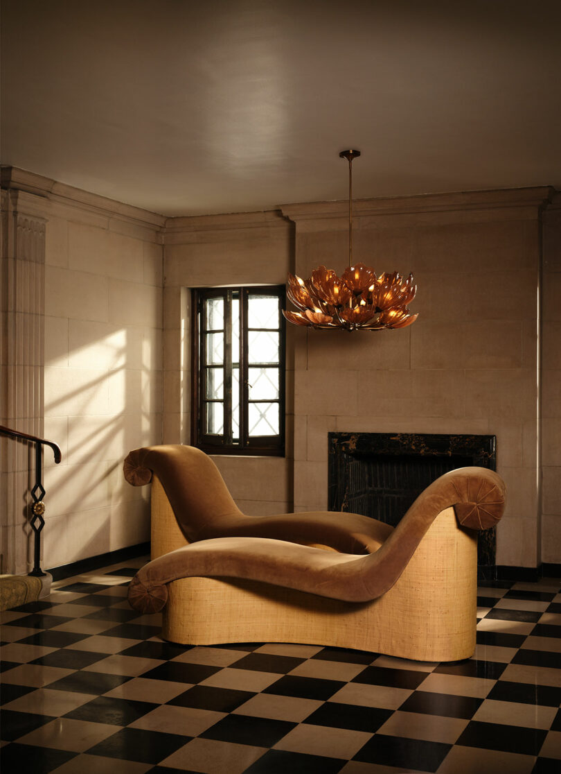 A beige, curved chaise lounge sits on a black-and-white checkered floor beneath a bronze floral chandelier in a room with stone walls and a window.