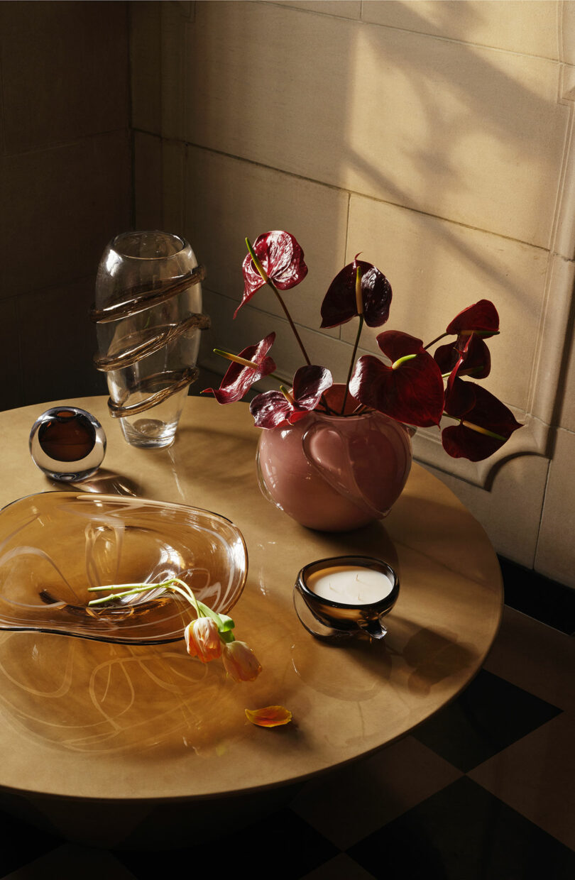 A round beige table with a pink vase of dark anthurium flowers, a glass sculpture, a glass bowl with orange tulips, and two round decorative objects, in sunlight.