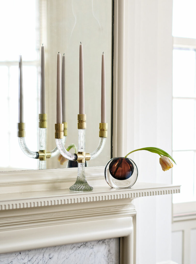 A glass and brass candelabra with three tall candles and a round vase with a single drooping tulip sit on a white mantel in front of a large mirror.