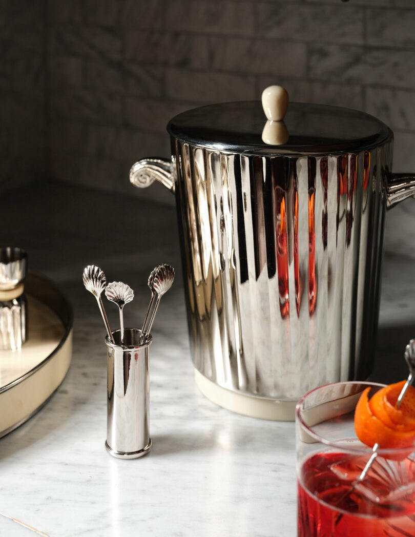 A polished metal ice bucket with lid, a small utensil holder with decorative stirrers, and two cocktails with orange garnish on a marble countertop.