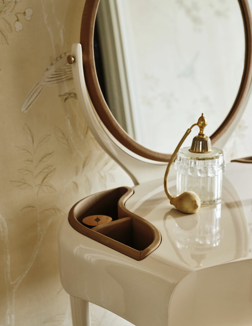 A close-up of a cream-colored vanity table with a round mirror, a vintage perfume atomizer, and small storage compartments.