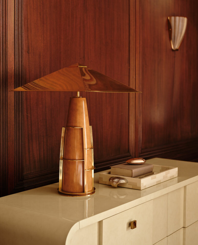 A brass and wood table lamp with a wide, conical shade sits on a cream-colored cabinet against a wood-paneled wall. A tray with a remote and a decorative object is beside it.