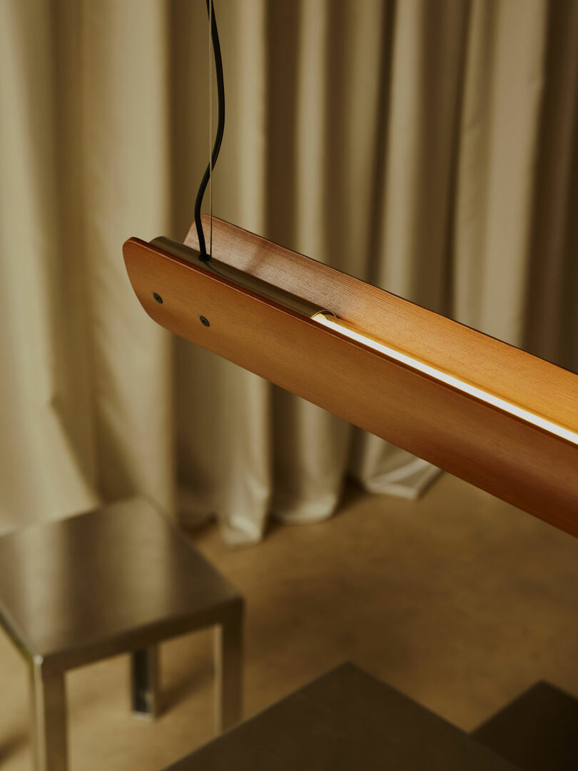 A wooden pendant light fixture with a visible black cord hangs above a metallic chair and table, with beige curtains in the background.