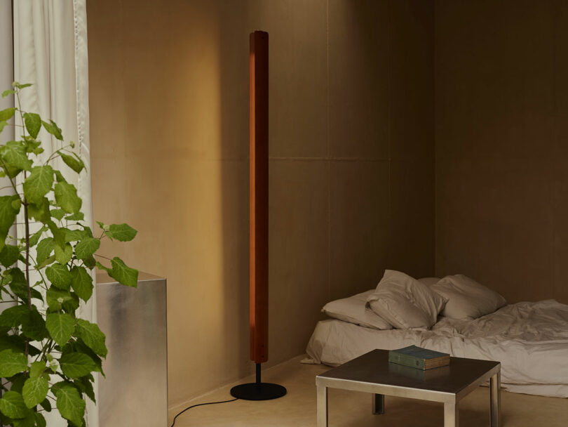 A minimal bedroom with beige walls, a floor lamp, unmade bed with white bedding, metal table with books, and a leafy green plant by the window.