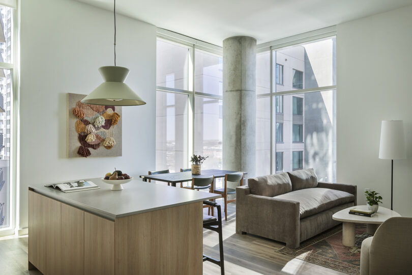 Modern Roost Rainey apartment living area with large windows, a small kitchen island, dining table, beige sofa, and wall art. Natural light fills the space, highlighting minimalist decor.