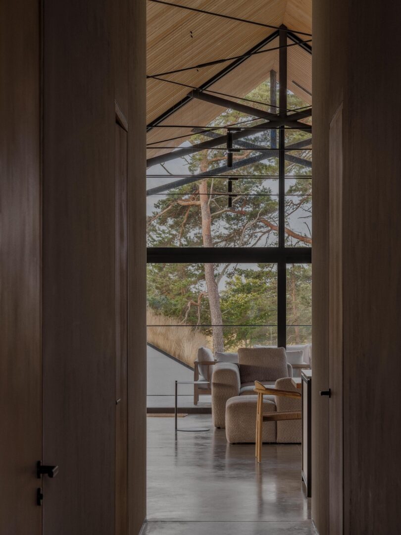 View through a hallway into a living area with beige furniture, large floor-to-ceiling windows, a high wooden ceiling, and a scenic forest visible outside.
