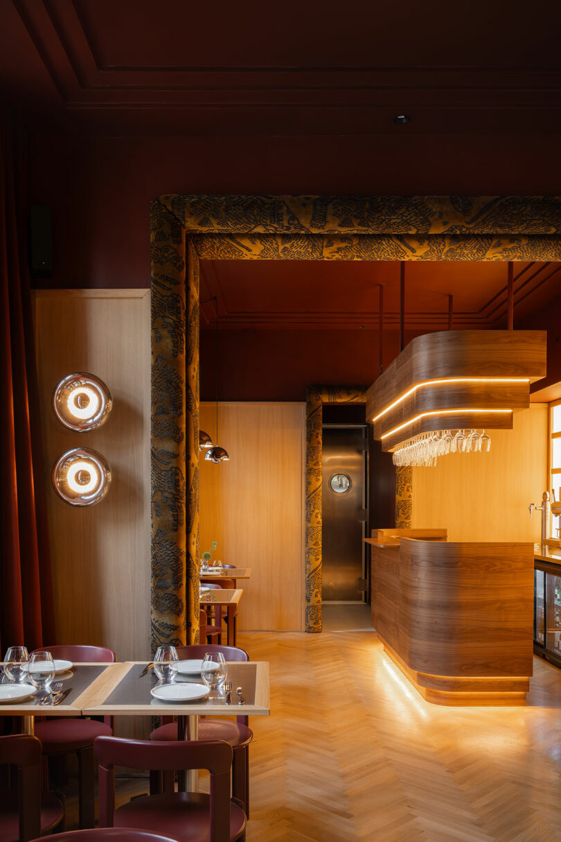 Modern restaurant interior with wood accents, ambient lighting, set tables with glassware, and a bar area with hanging glasses.