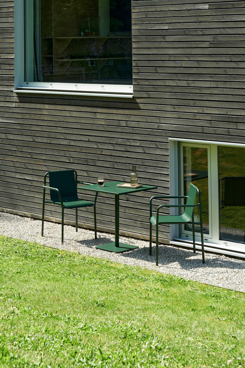 Two green chairs and a green table with drinks invite you to tiptoe across the gravel outside a modern wooden building, nestled beside a grassy lawn and large windows.