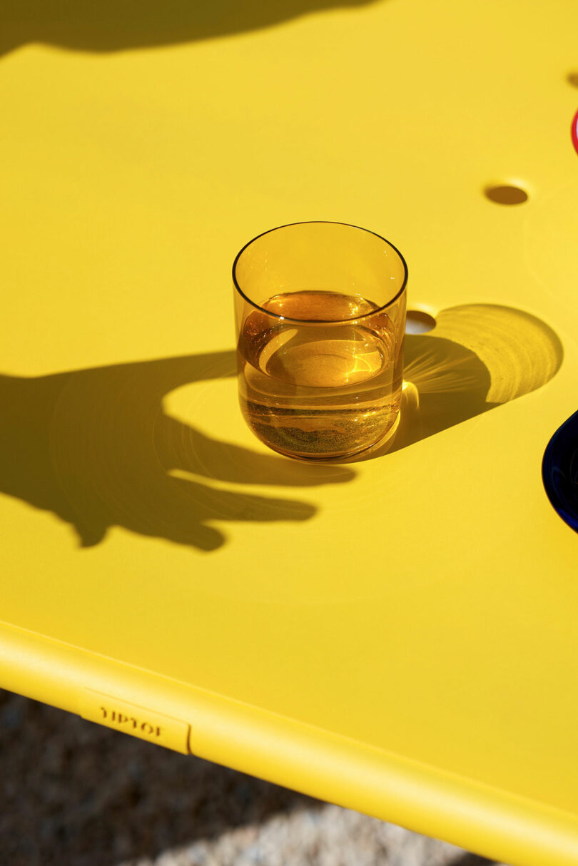A transparent amber glass of liquid sits on a bright yellow table, casting a shadow. A hand’s shadow seems to tiptoe toward the glass, adding a sense of anticipation to the scene.