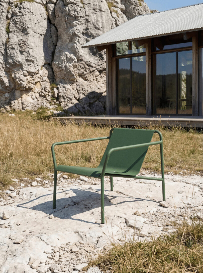 A green metal outdoor chair sits on a stone path in front of a modern building with large windows, set in a grassy area where you can tiptoe near a rocky cliff.