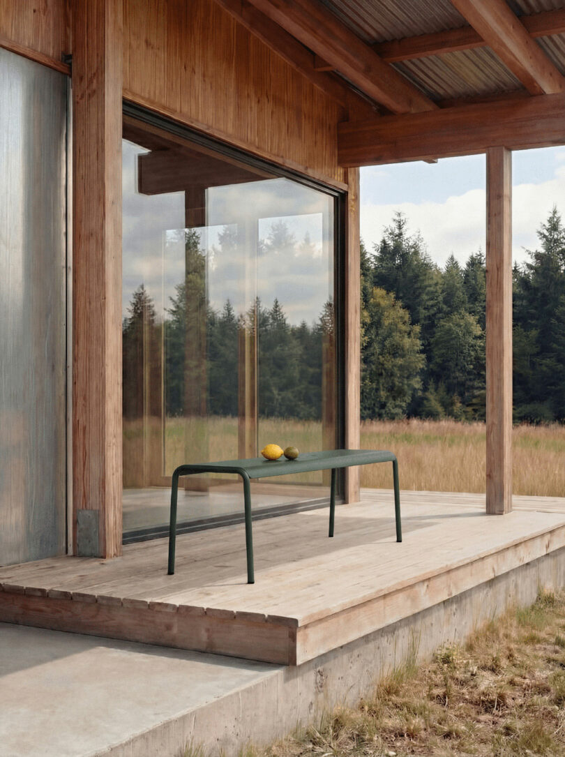 A rectangular green table with two yellow fruits sits on a wooden porch of a modern house, as children tiptoe nearby, surrounded by grass and trees under a partly cloudy sky.