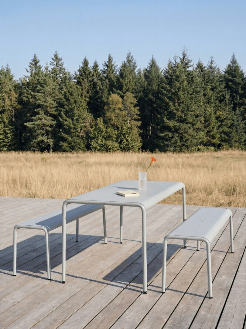 Outdoor metal table with two benches on a wooden deck, a clear vase with a single orange flower tiptoe across the table, and trees in the background.