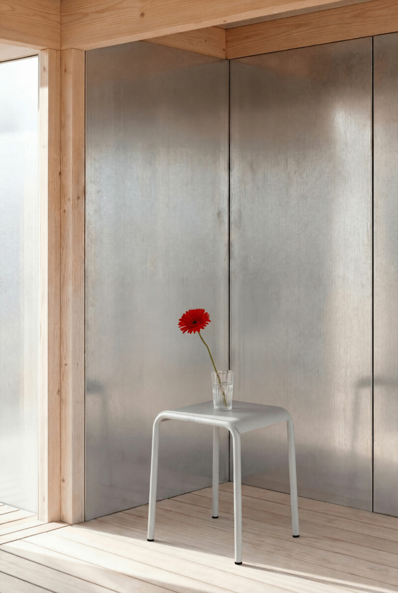 A single red flower in a glass of water sits tiptoe on a small white stool in a minimalist, sunlit room with metallic walls and wooden beams.