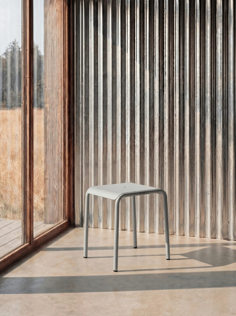 A simple gray metal stool stands on a concrete floor near glass walls and a corrugated metal panel, with sunlight casting shadows inside the room—as if inviting you to tiptoe closer and enjoy the quiet ambiance.