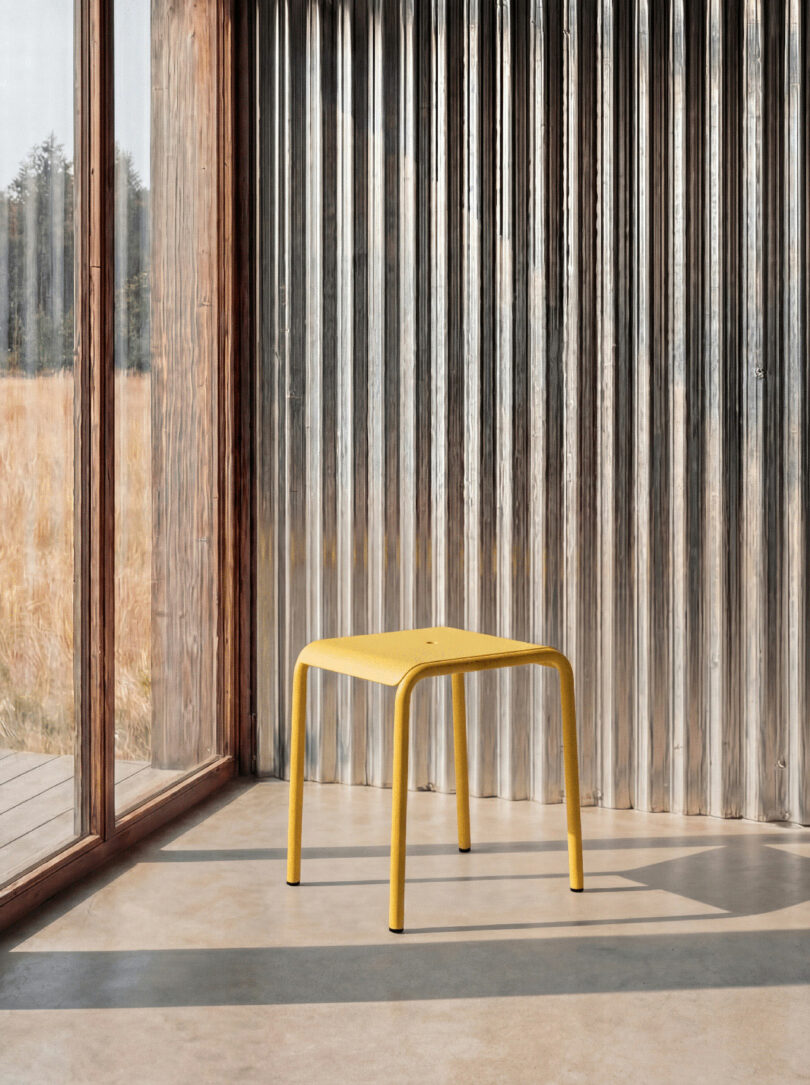 A yellow Tiptoe stool with rounded legs sits on a concrete floor next to a large window and corrugated metal wall, with outdoor grass visible through the glass.
