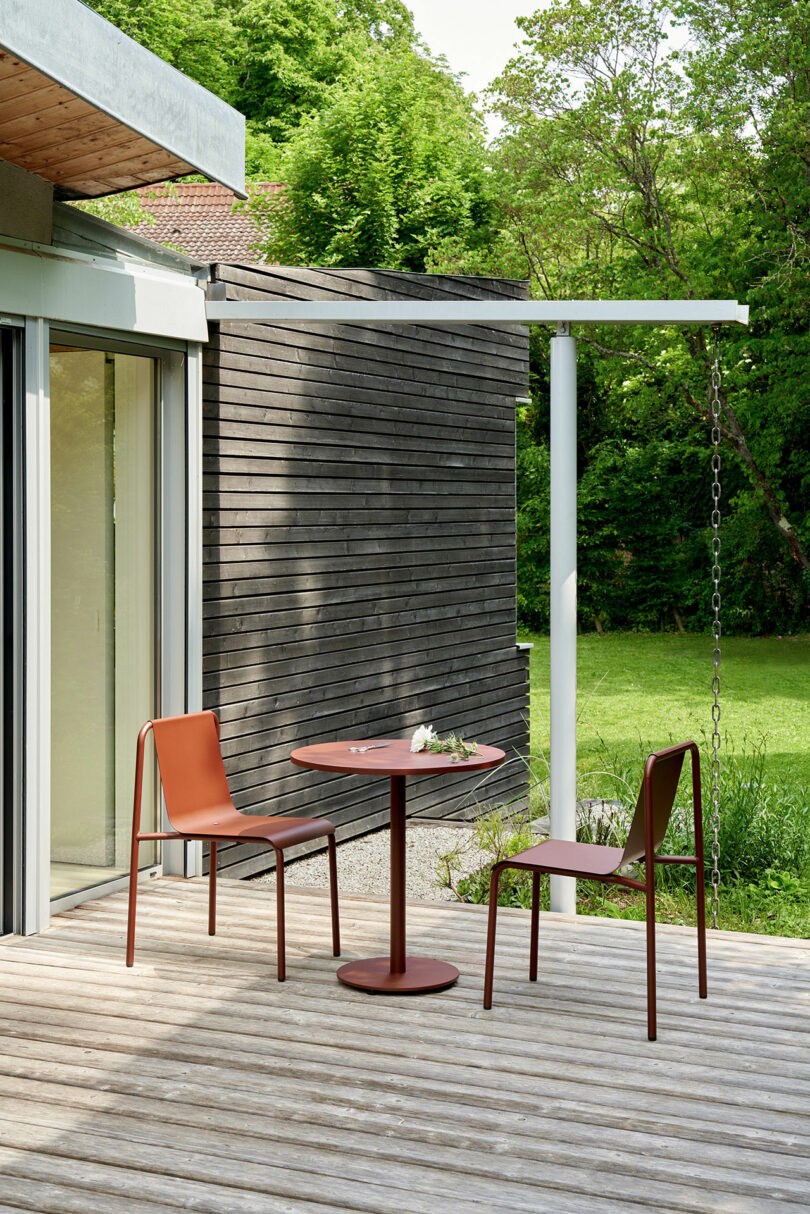 A small round table with two brown chairs sits on a wooden deck outside a modern house, inviting you to tiptoe outside and enjoy the greenery and trees in the background.