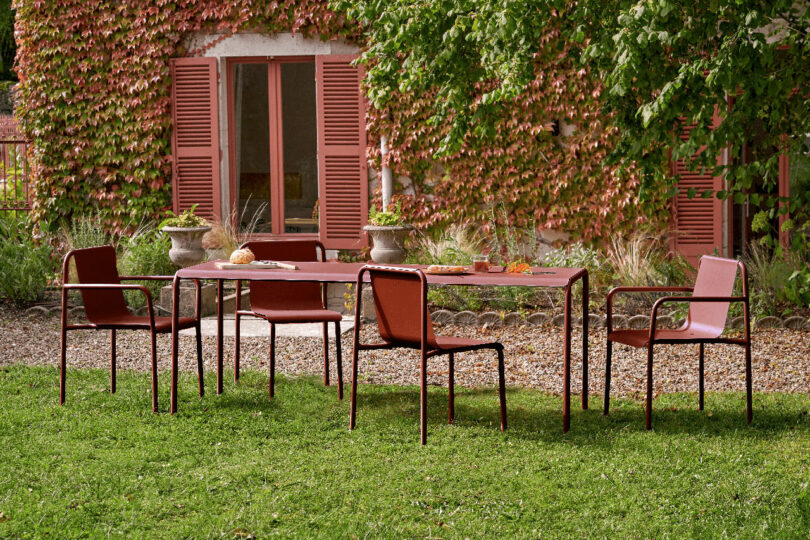 A rectangular outdoor table with five red chairs stands on grass near a house with red shutters, surrounded by greenery and vines that seem to tiptoe up the walls.