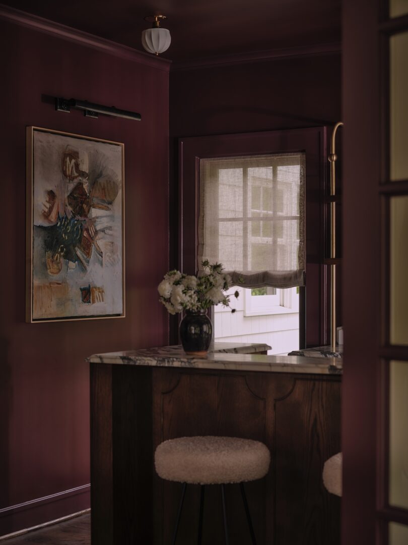A dimly lit room with purple walls, abstract art, a marble counter with flowers in a vase, two cushioned stools, and a window with sheer curtains.