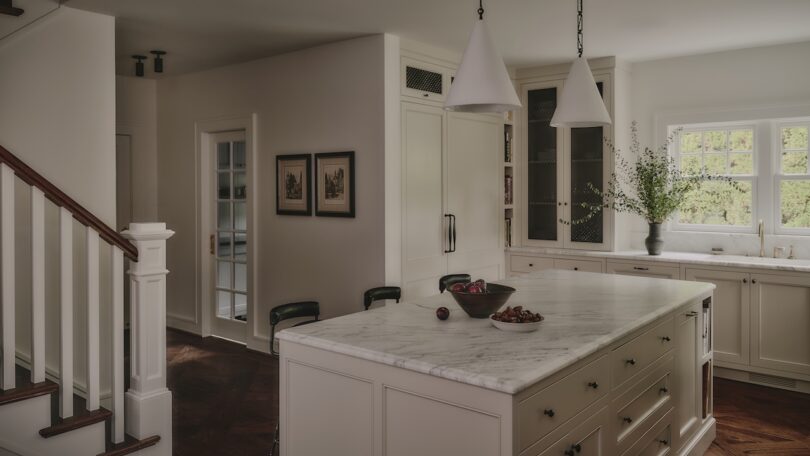 A modern kitchen with a large marble island, white cabinets, pendant lights, barstools, fruit bowls, and a window above the sink letting in natural light.