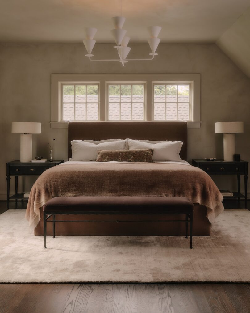 A neatly made bed with brown and beige bedding, flanked by two black nightstands with lamps, sits below four windows in a softly lit bedroom. A bench is at the foot of the bed.