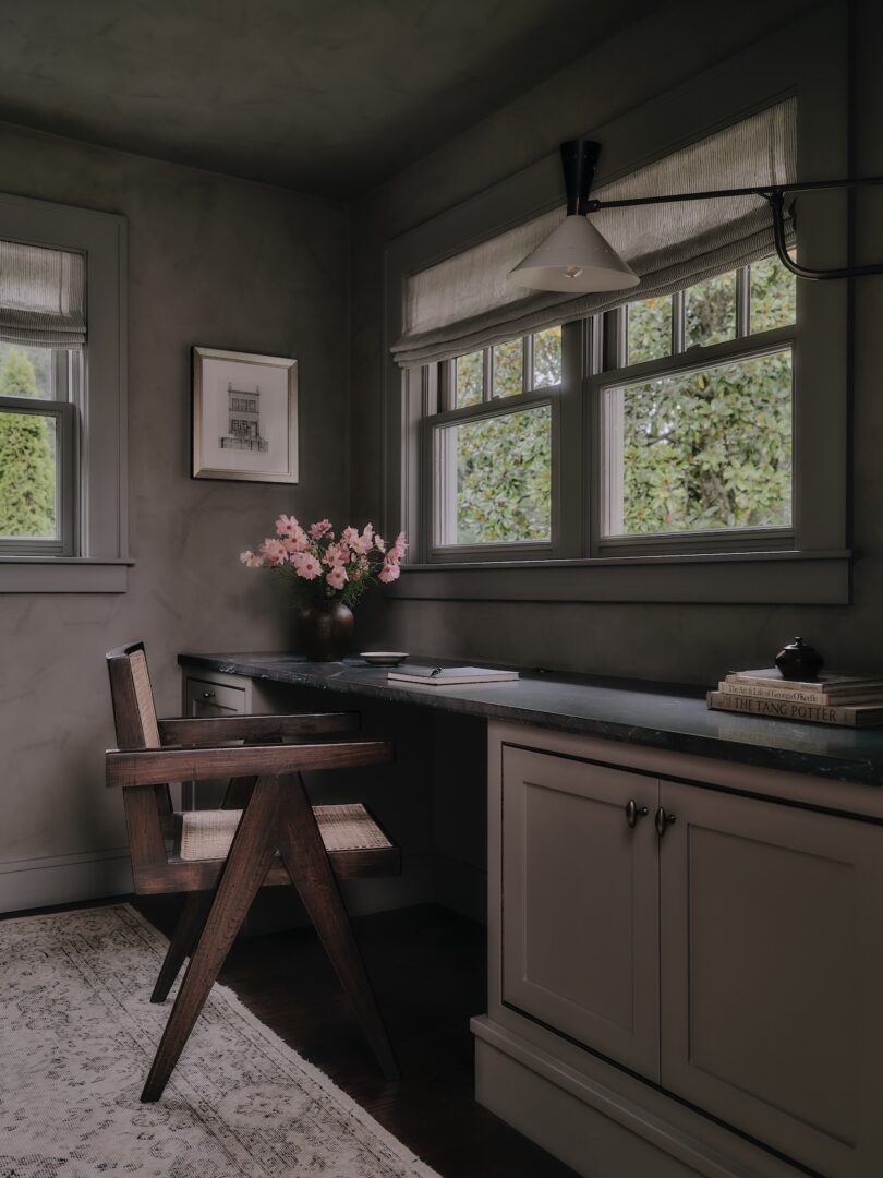 A home office with a wooden chair, built-in desk, books, pink flowers in a vase, framed art, and two windows with Roman shades overlooking greenery.