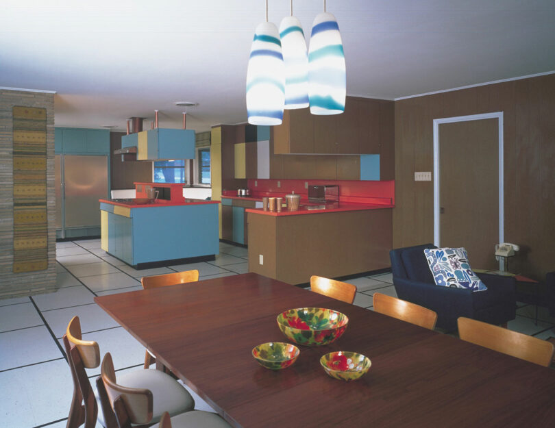 Mid-century modern kitchen and dining area with blue and brown cabinetry, red countertops, pendant lights, and wooden dining table with colorful glass bowls.