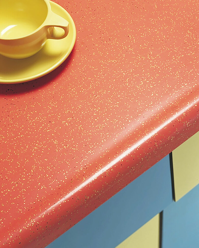 A yellow cup and saucer on a red speckled countertop with blue and yellow cabinets underneath.