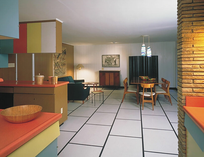Mid-century modern living and dining area with colorful cabinets, wood furniture, and geometric floor tiles; a stone wall is visible on the right.