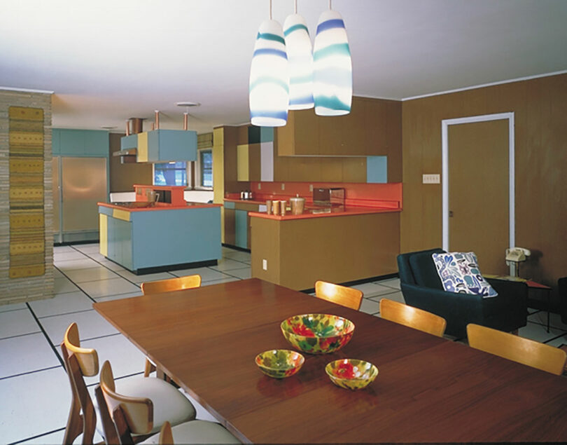 Mid-century modern kitchen and dining area with pastel cabinets, red countertops, wooden furniture, striped pendant lights, and decorative bowls on the table.