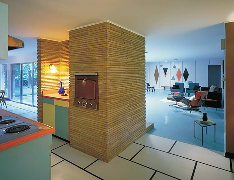 Mid-century modern interior with a central brick pillar, built-in oven, pastel cabinetry, open living area, and geometric wall art. Large windows provide natural light.