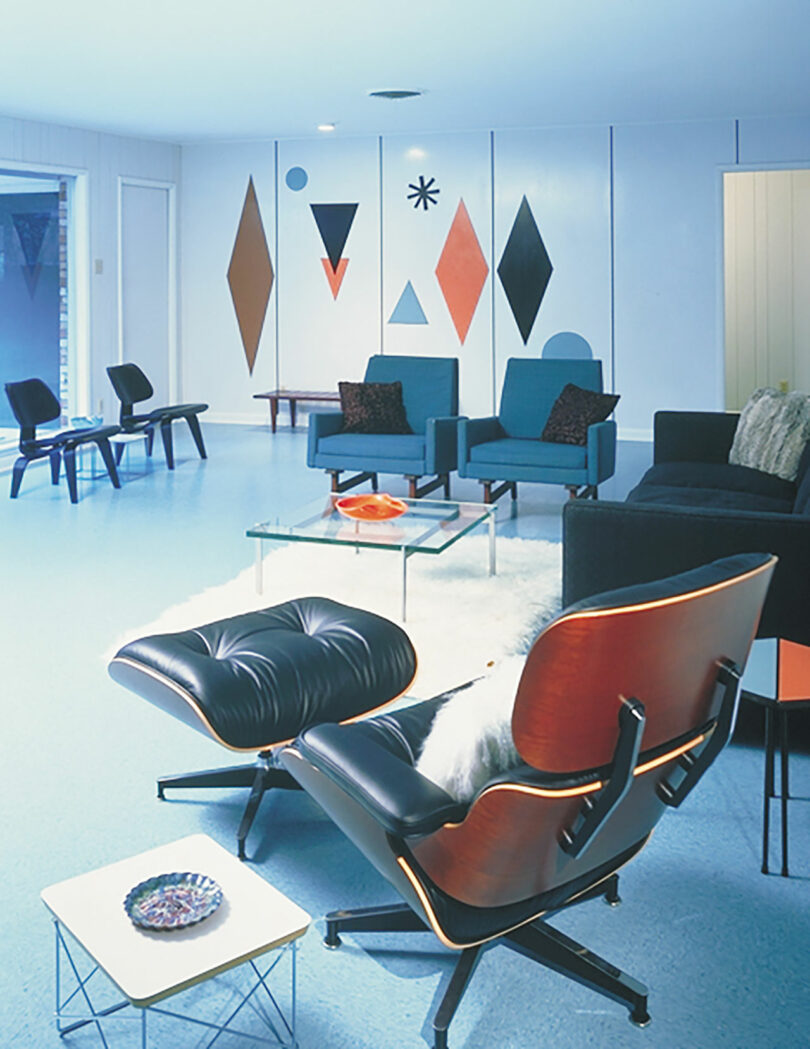 Mid-century modern living room with blue flooring, black and blue furniture, a glass coffee table, and geometric wall art in orange, black, and brown shapes.