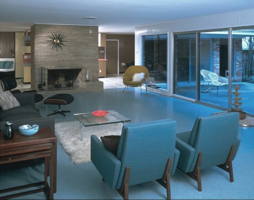 A mid-century modern living room with blue chairs, a fireplace, glass coffee table, and sliding doors opening to an outdoor patio with wire chairs.