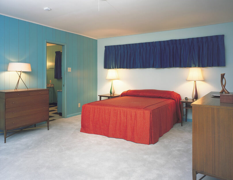 Bedroom with light blue walls, red bedspread, two bedside tables with lamps, blue curtains, dresser, and carpeted floor.