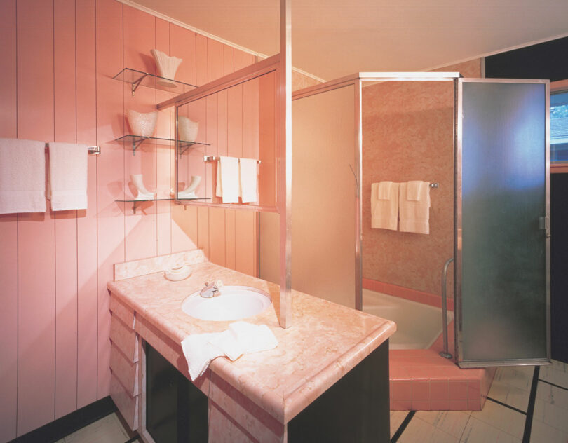 A bathroom with pink wall panels, a pink marble countertop, a sink, shelves with white decor, and a shower with frosted glass doors and white towels.