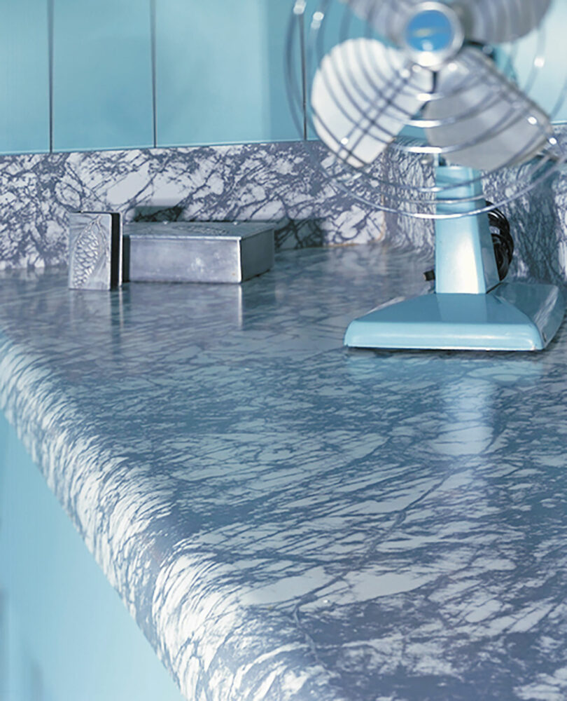 Close-up of a blue and white marbled countertop with a metal box and a vintage blue fan in a retro-styled room.