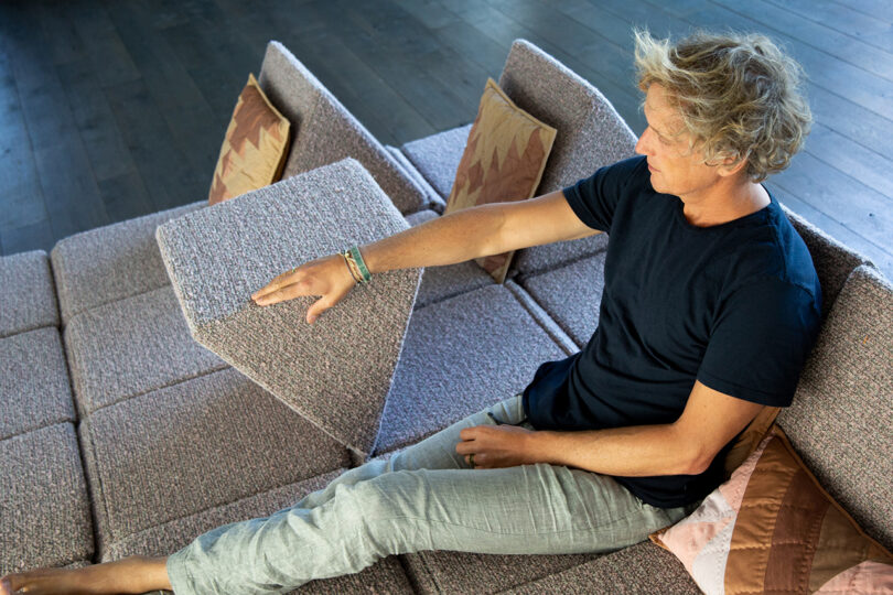 A person sitting on a modular sofa lifts one of the adjustable backrests, with pillows and a wooden floor visible in the background.
