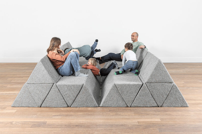 Four adults and two children sit and play on a modular gray geometric sofa in a minimalist room with light wood flooring and a white wall.