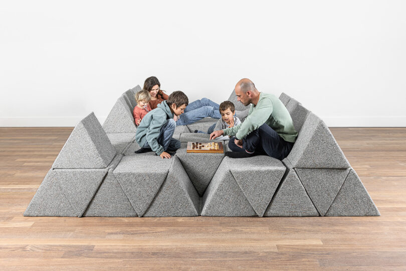 A family of five sits on a modular, geometric gray sofa, playing chess together on a wooden floor against a plain white wall.