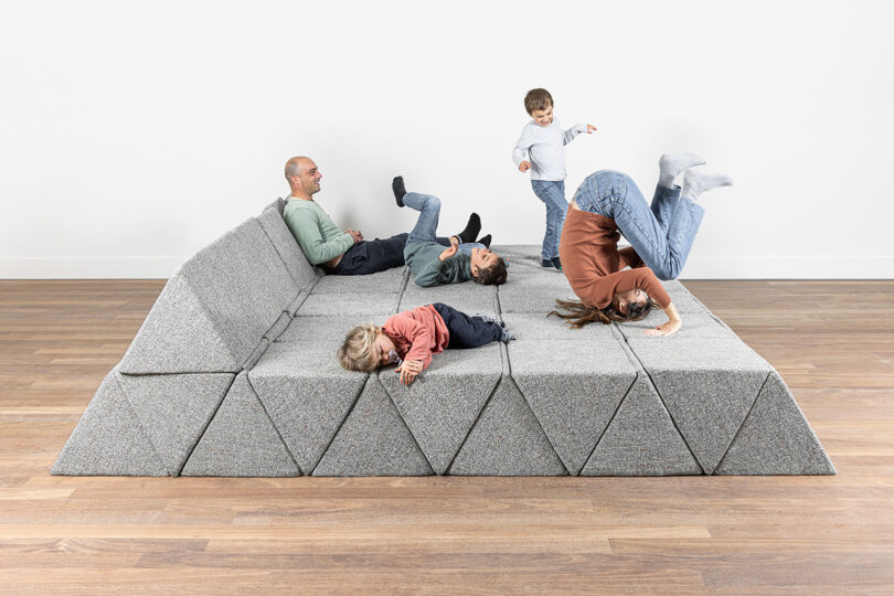 An adult and four children play and relax on a large modular gray cushioned seating platform in a bright room with wooden floors.
