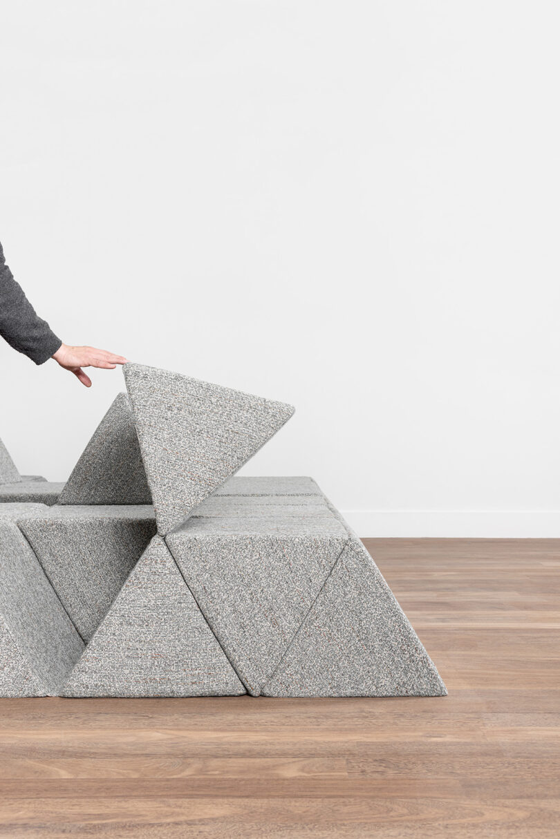 A person lifts a triangular gray cushion from a modular arrangement of similar cushions on a wooden floor against a plain white wall.
