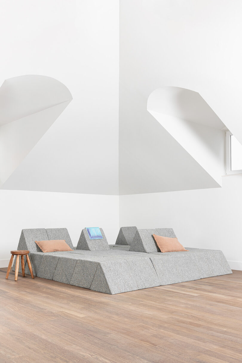 A minimalist room with angular gray modular seating, two tan cushions, a wooden stool, and white walls with geometric ceiling details.